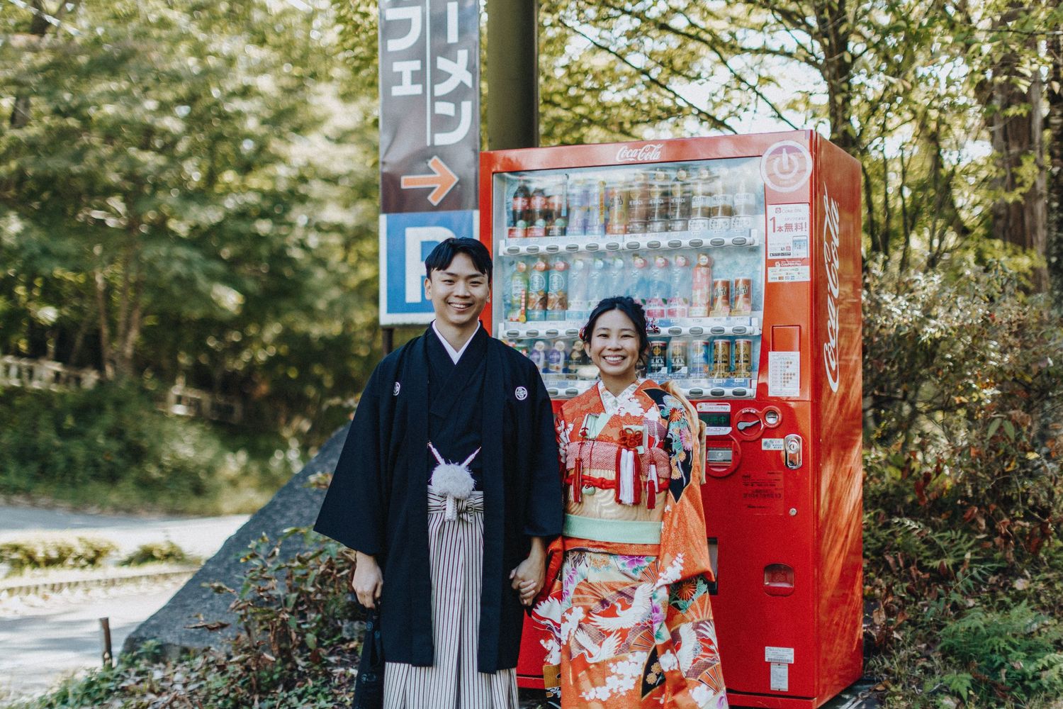 Two people pose next to a vintage red Coca-Cola vending machine in an outdoor garden setting.