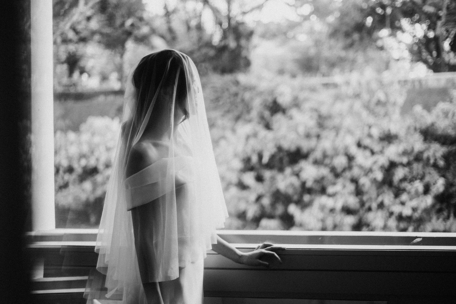 A bride in a flowing white veil stands silhouetted against a window overlooking a garden in black and white photography.