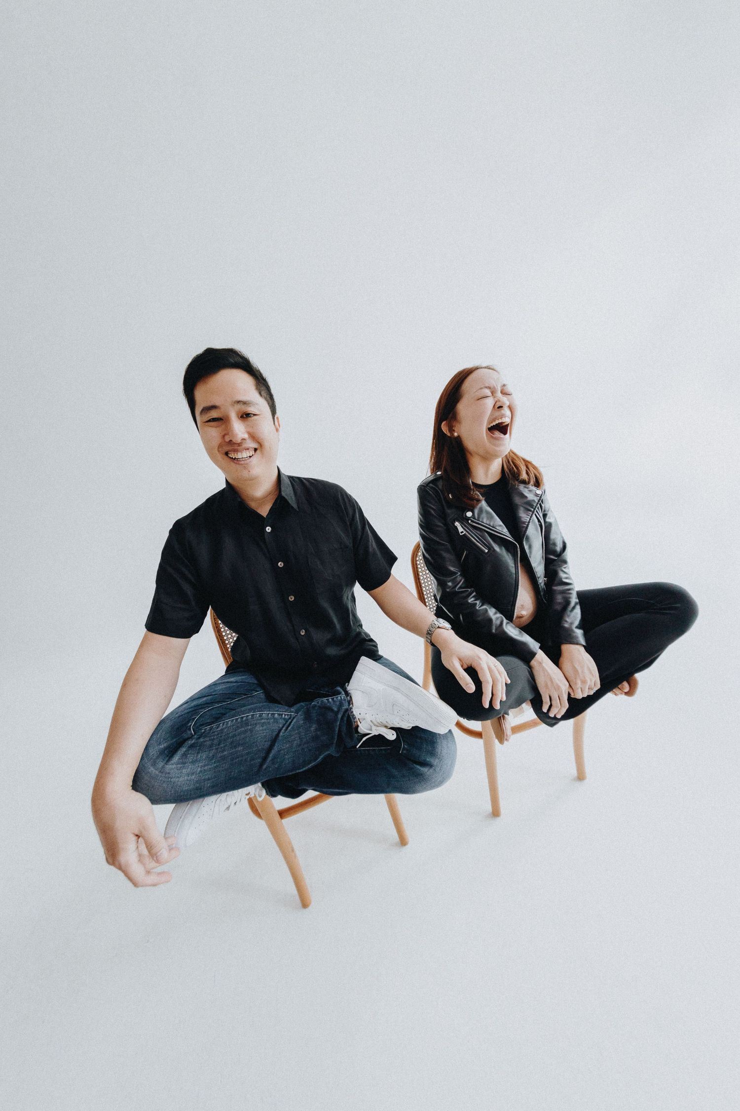 Candid studio shot of two people sitting and laughing on wooden chairs against white background.