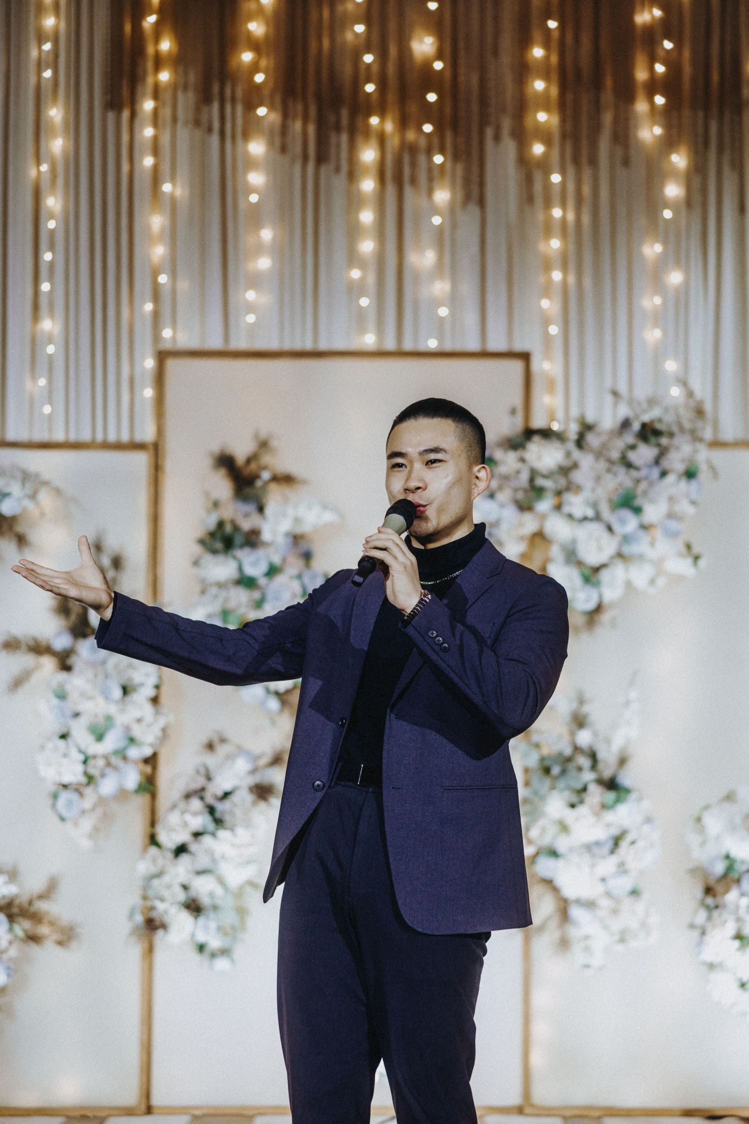 A performer in a navy suit sings with a microphone against a backdrop of fairy lights and white floral decorations.