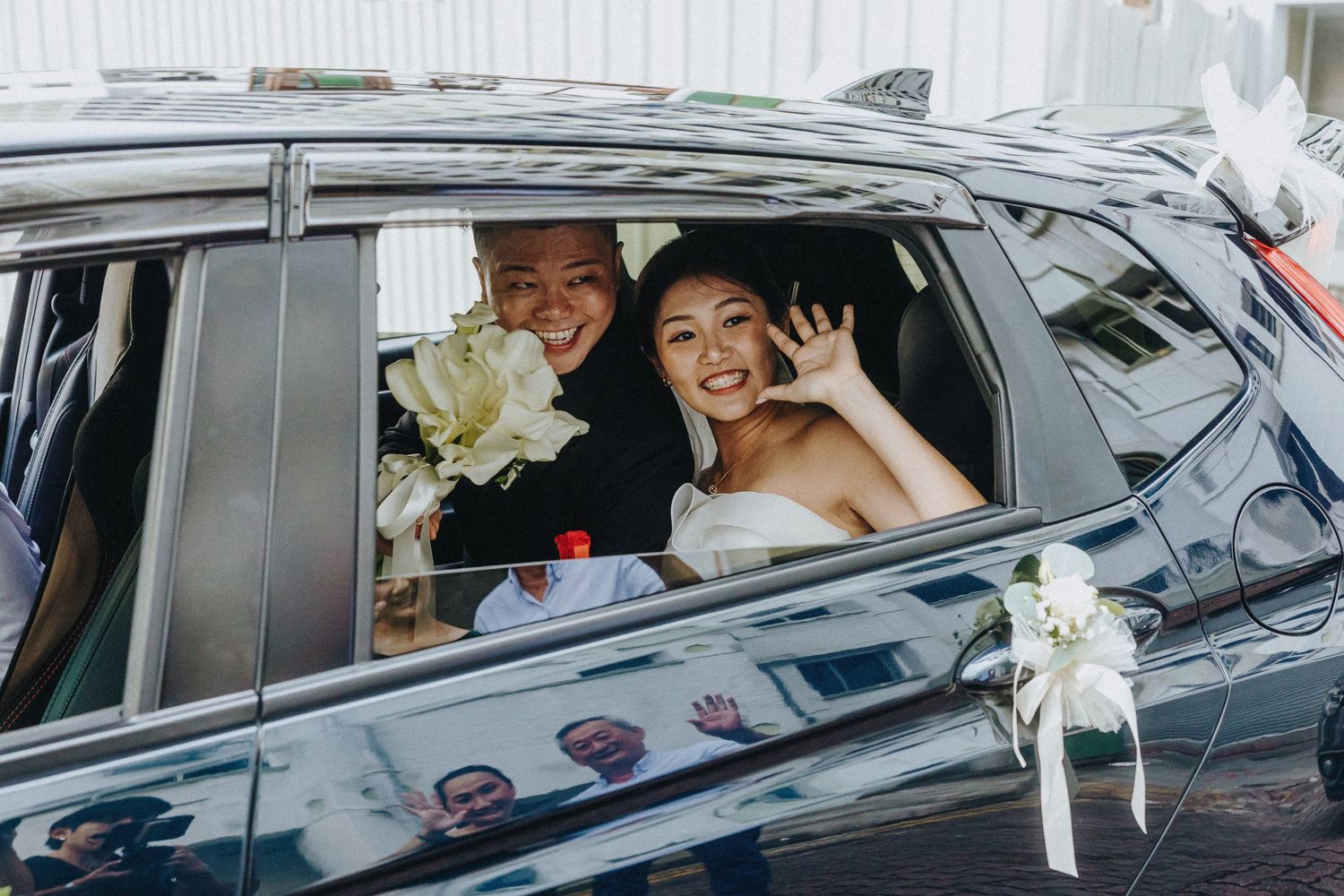 Newlyweds waving from inside a luxury car decorated with white flowers.