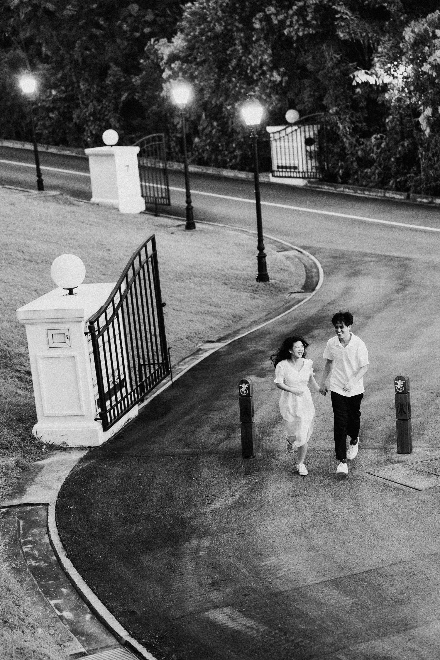 Couple walking along curved pathway with ornate lamp posts and iron gates at dusk.