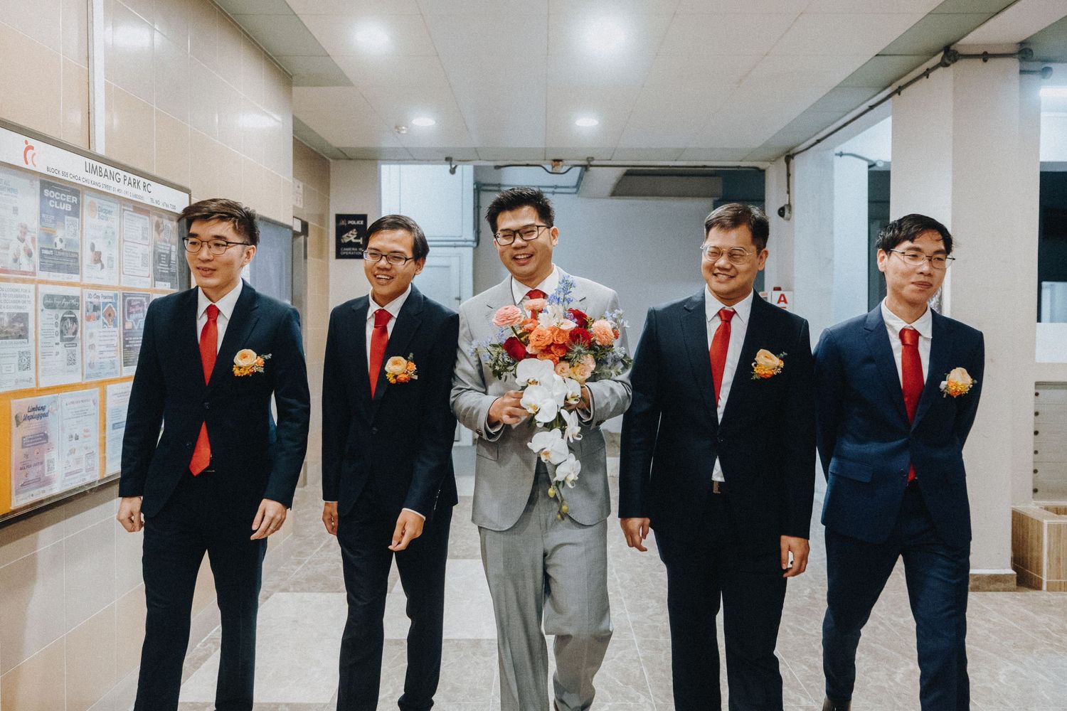 Wedding party members in black suits and red ties pose together in a bright indoor hallway.