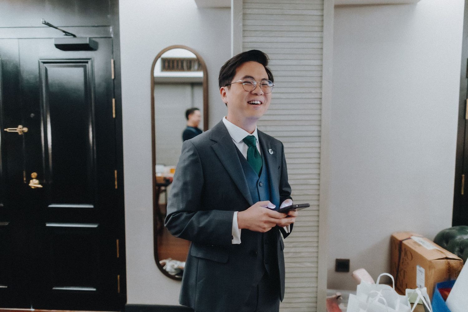 A person in a gray suit smiling and holding something while standing in front of a black door in an indoor setting.