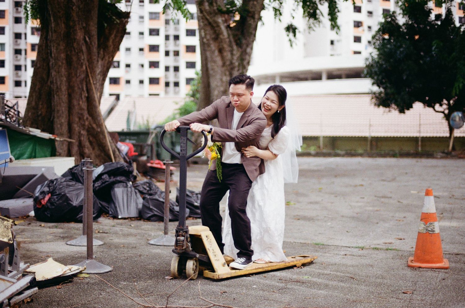 A couple shares a joyful moment while balancing on a wooden board outdoors near apartment buildings.