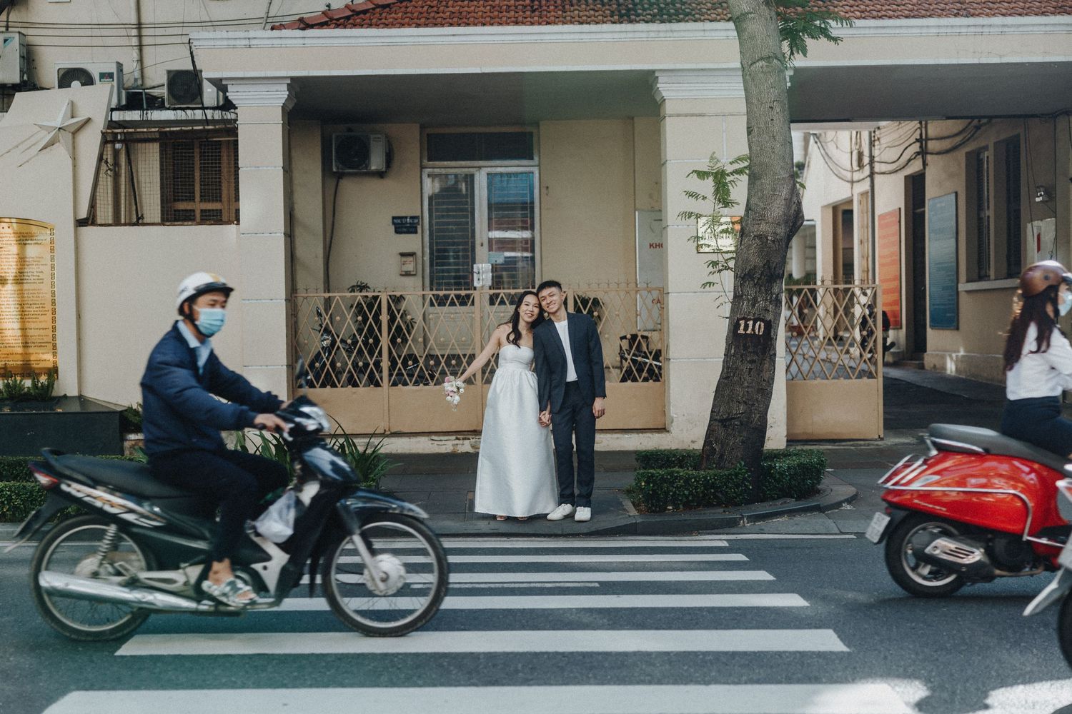 People stand on a city street corner with parked motorcycles and a red scooter near a pedestrian crossing.