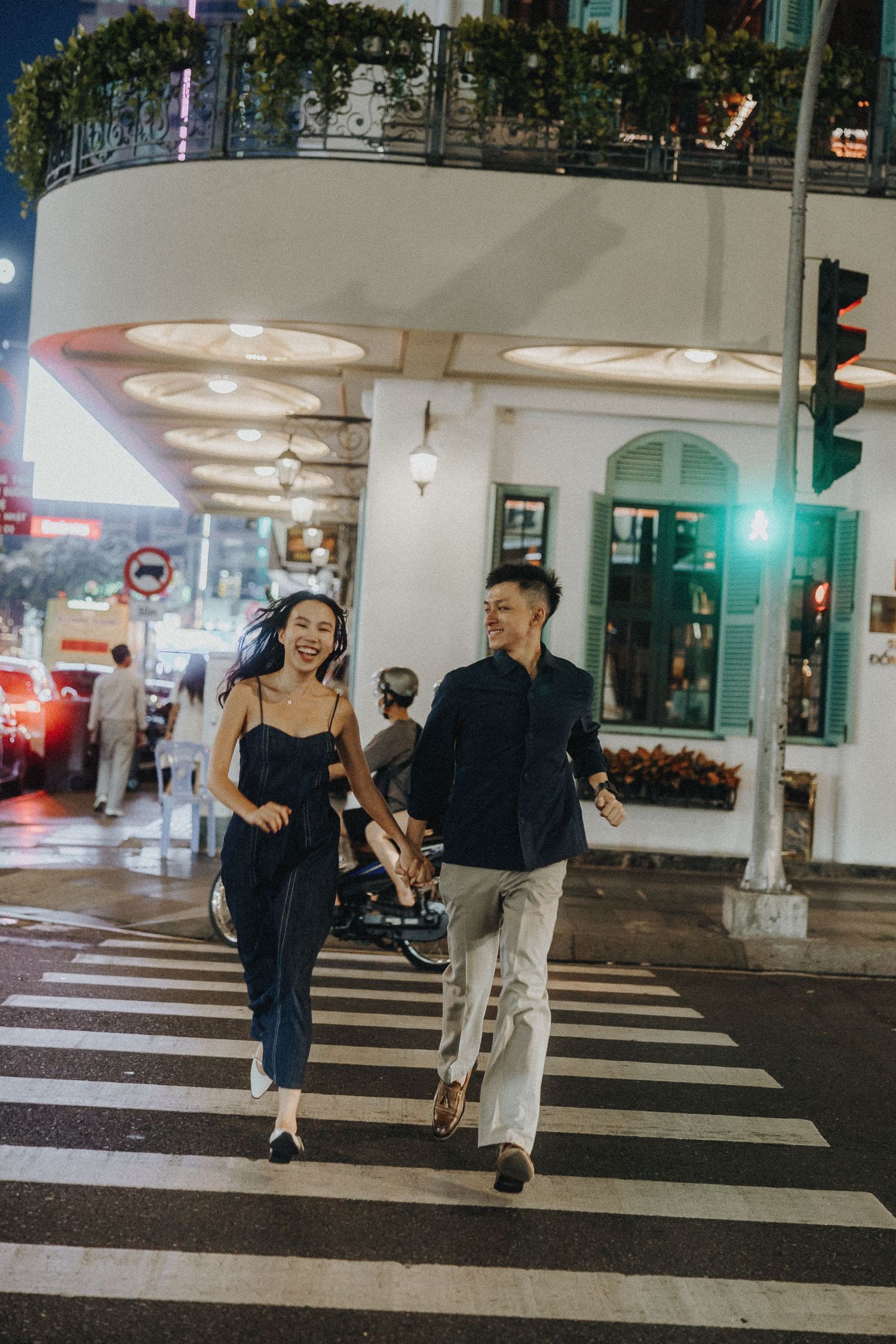 A couple strolls across an illuminated crosswalk at night in a stylish urban setting.