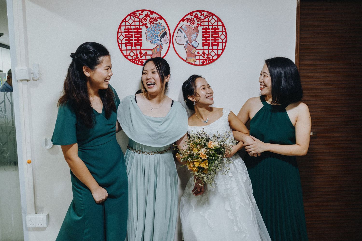 Bridal party in emerald green dresses sharing a candid laughing moment in front of traditional Chinese wall decorations.