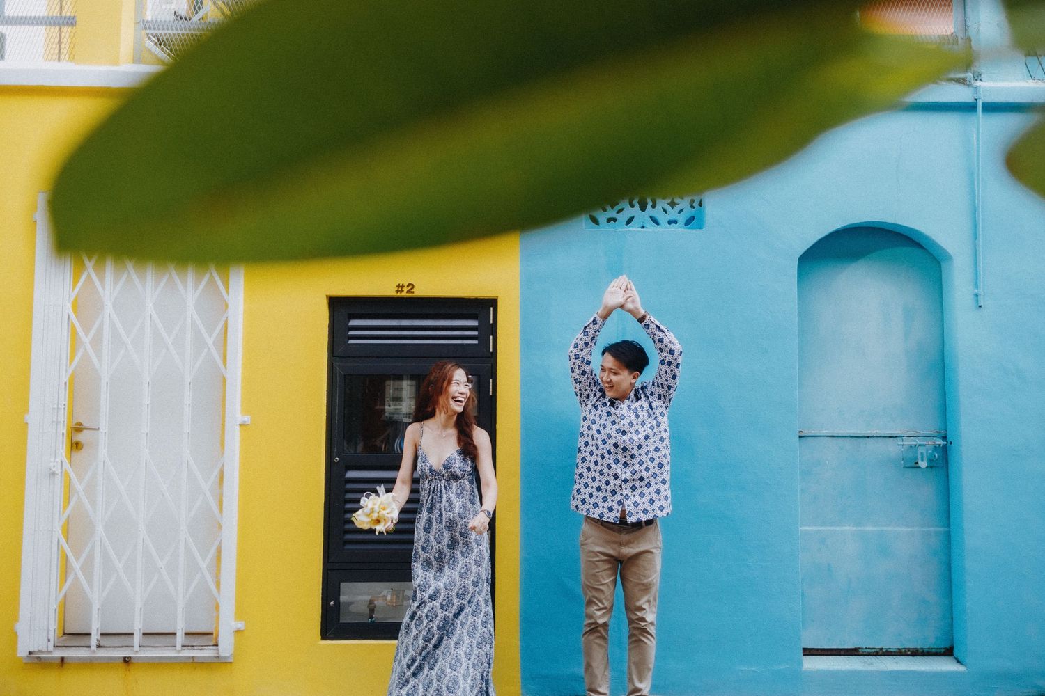 A playful couple poses against vibrant yellow and blue walls with contrasting architectural details.