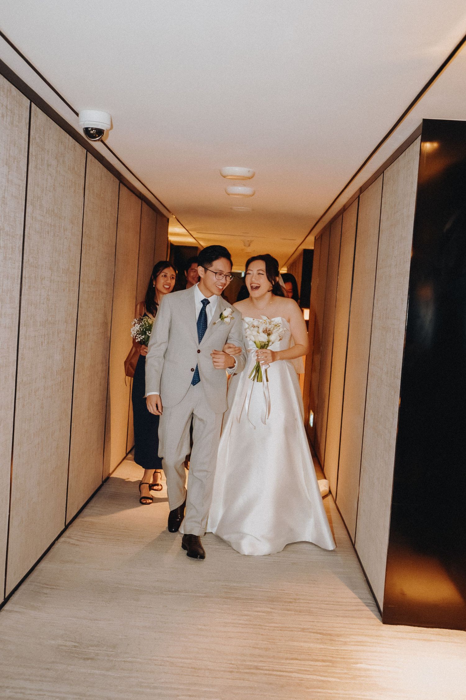 Wedding party members walk and dance down a modern hotel hallway.