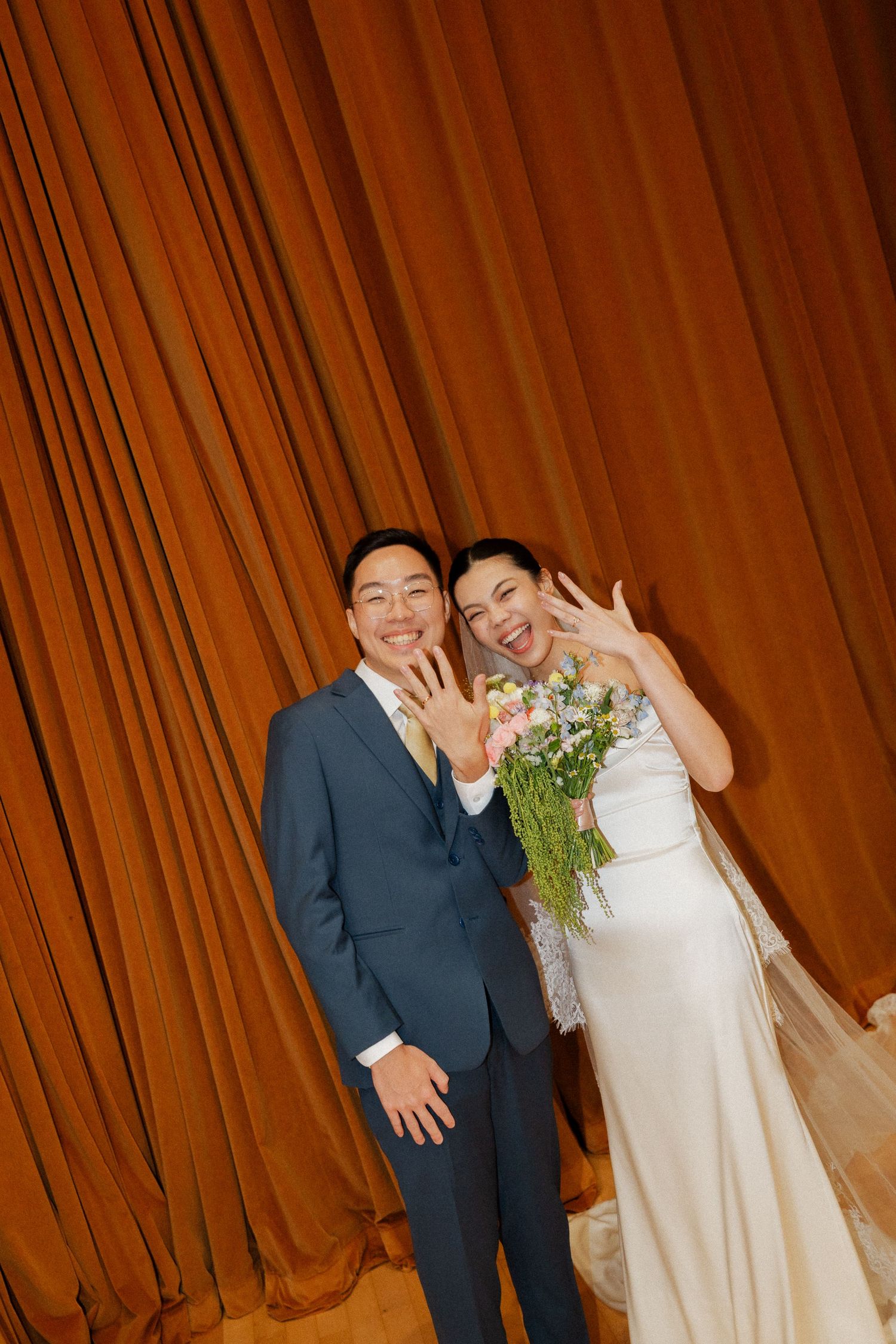 Newlyweds in blue suit and white gown showing wedding rings smiling before orange curtain.