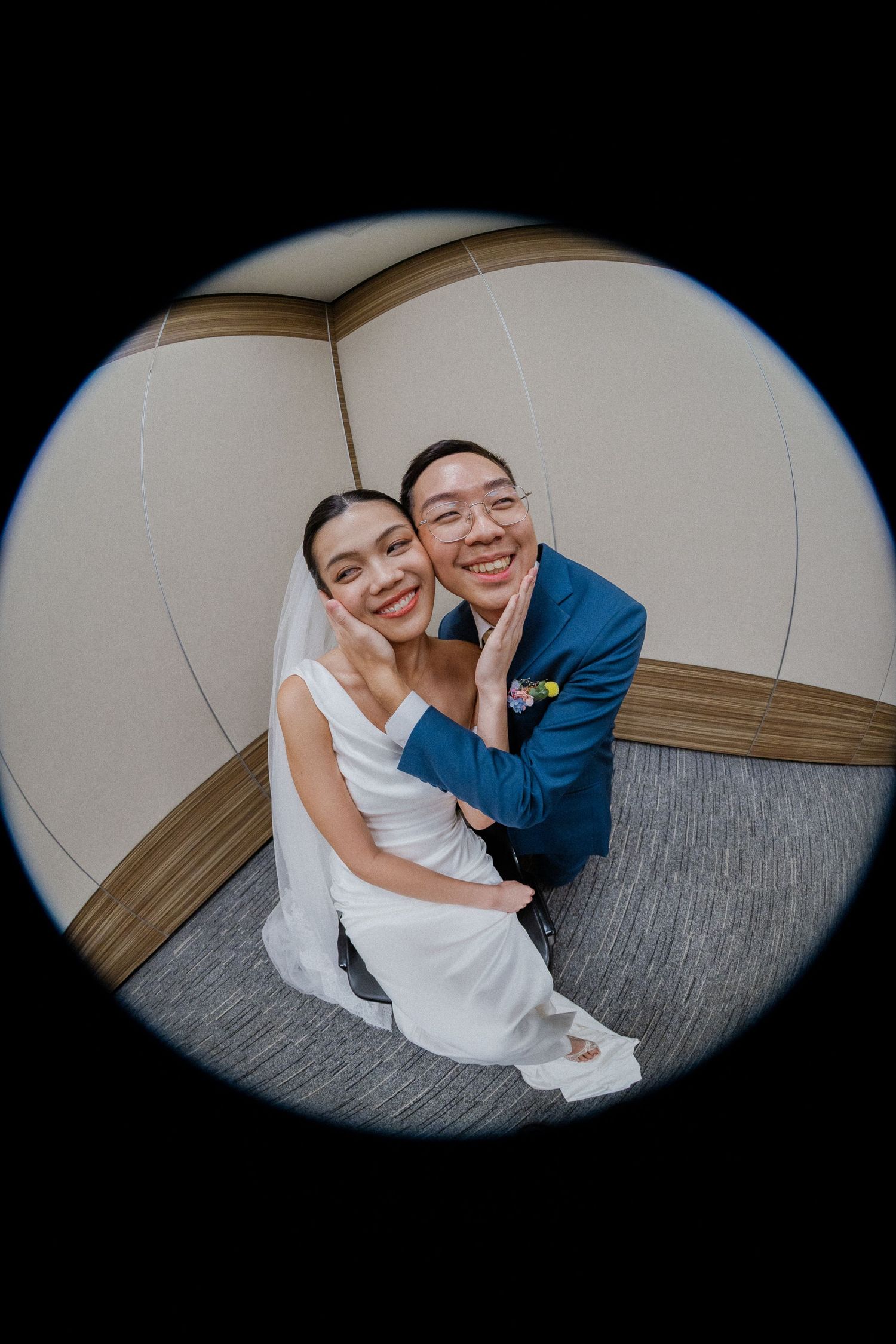 A joyful newlywed couple poses for a fisheye lens photo, bride in white gown and groom in blue suit, smiling warmly.