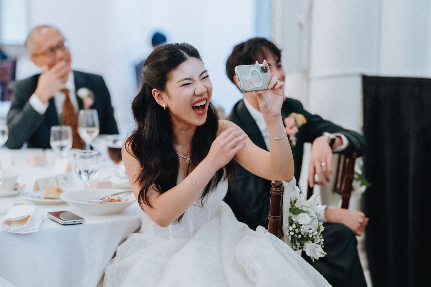 Bride laughing joyfully at wedding reception table while guest photographs moment.