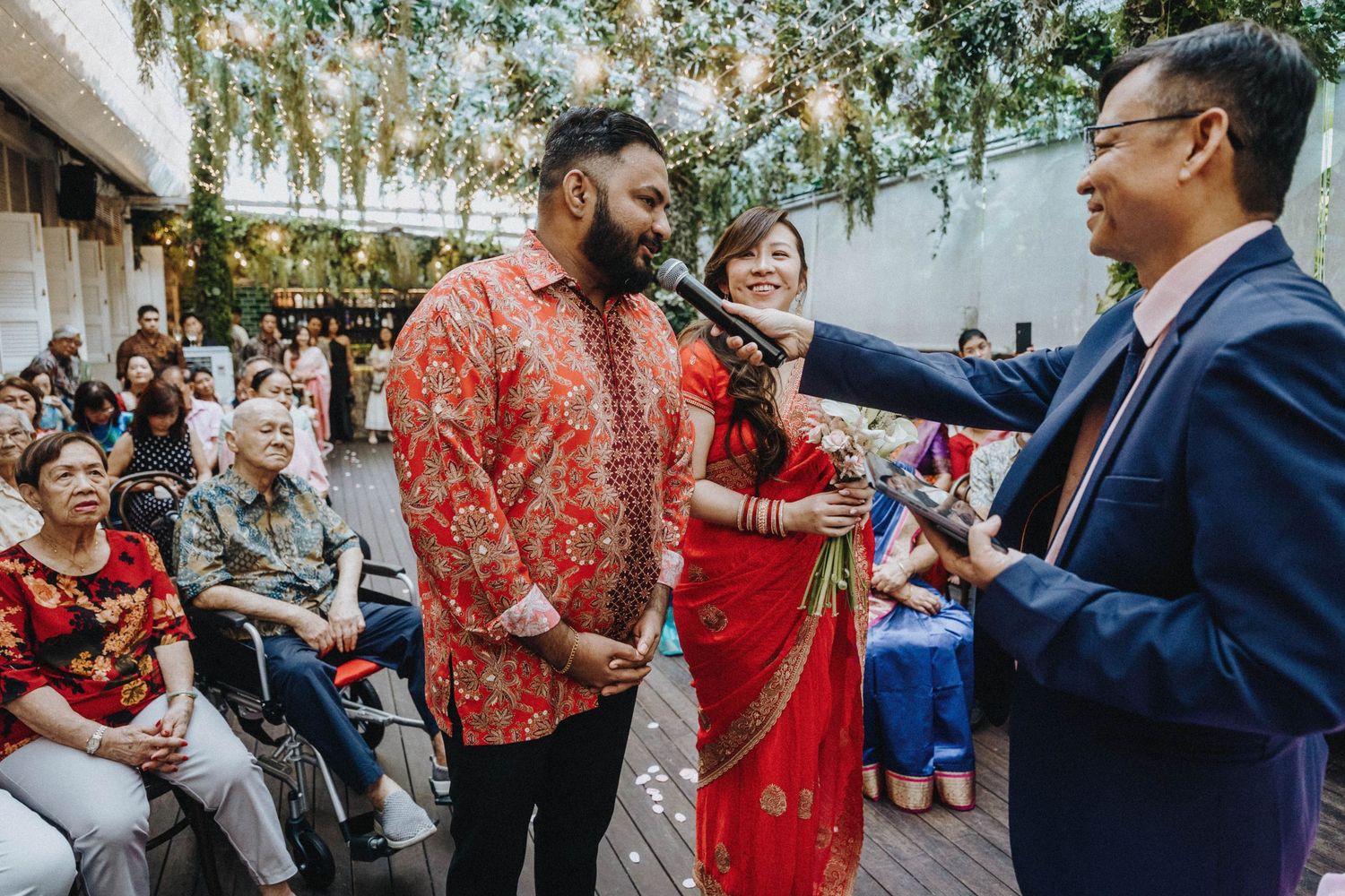 Couple in traditional Indian attire exchange vows at an outdoor wedding ceremony with seated guests.