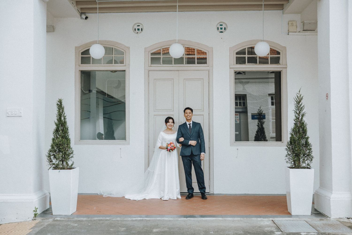 Asian wedding couple in formal attire standing before a white colonial building with arched windows and potted topiaries.