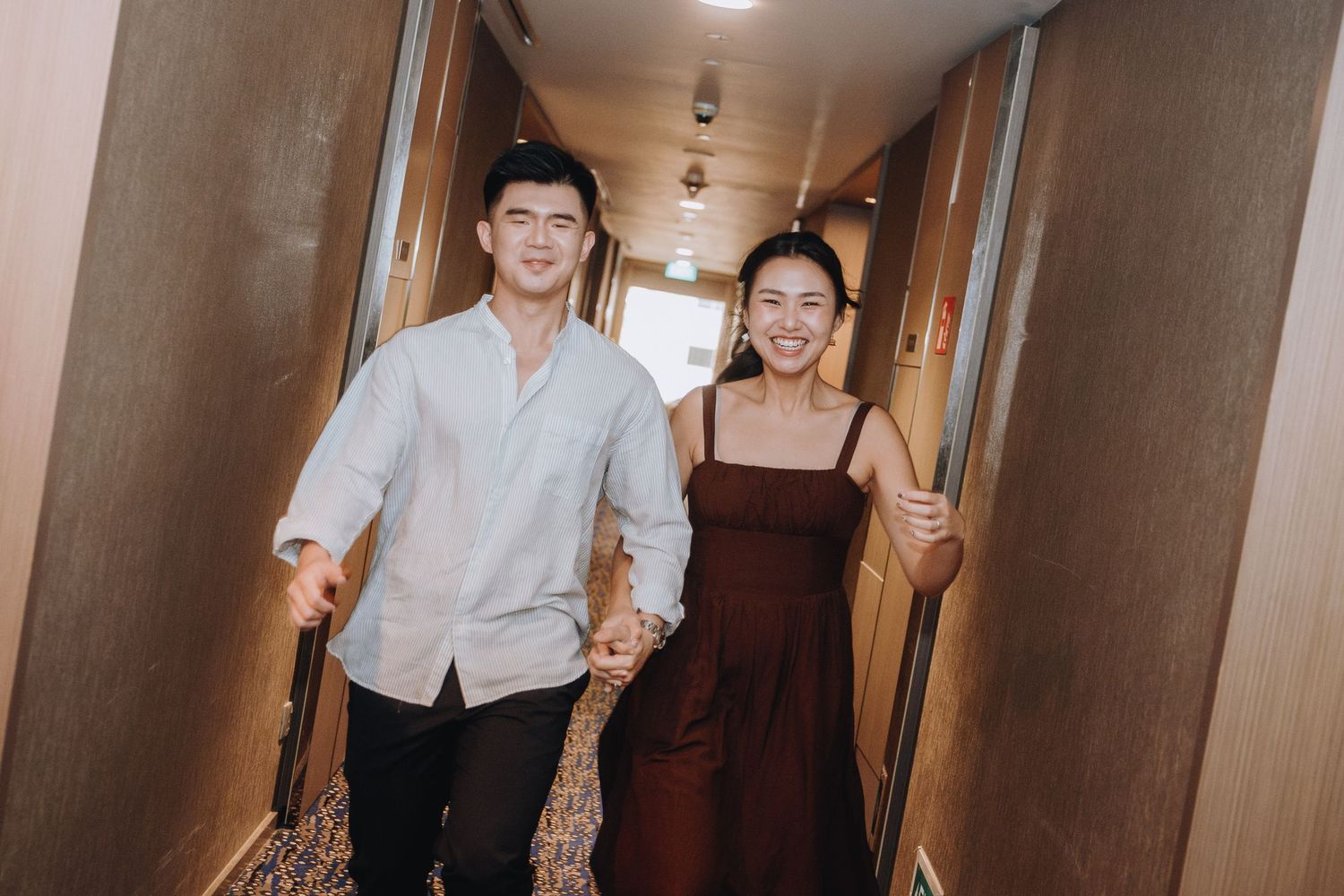 A smiling couple walks hand-in-hand down a hotel hallway, the man in a light shirt and woman in a burgundy dress.