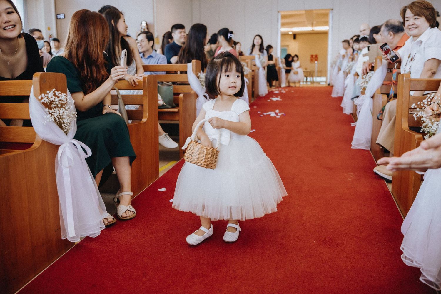 Adorable flower girl in white dress walks down red carpet aisle during wedding ceremony.