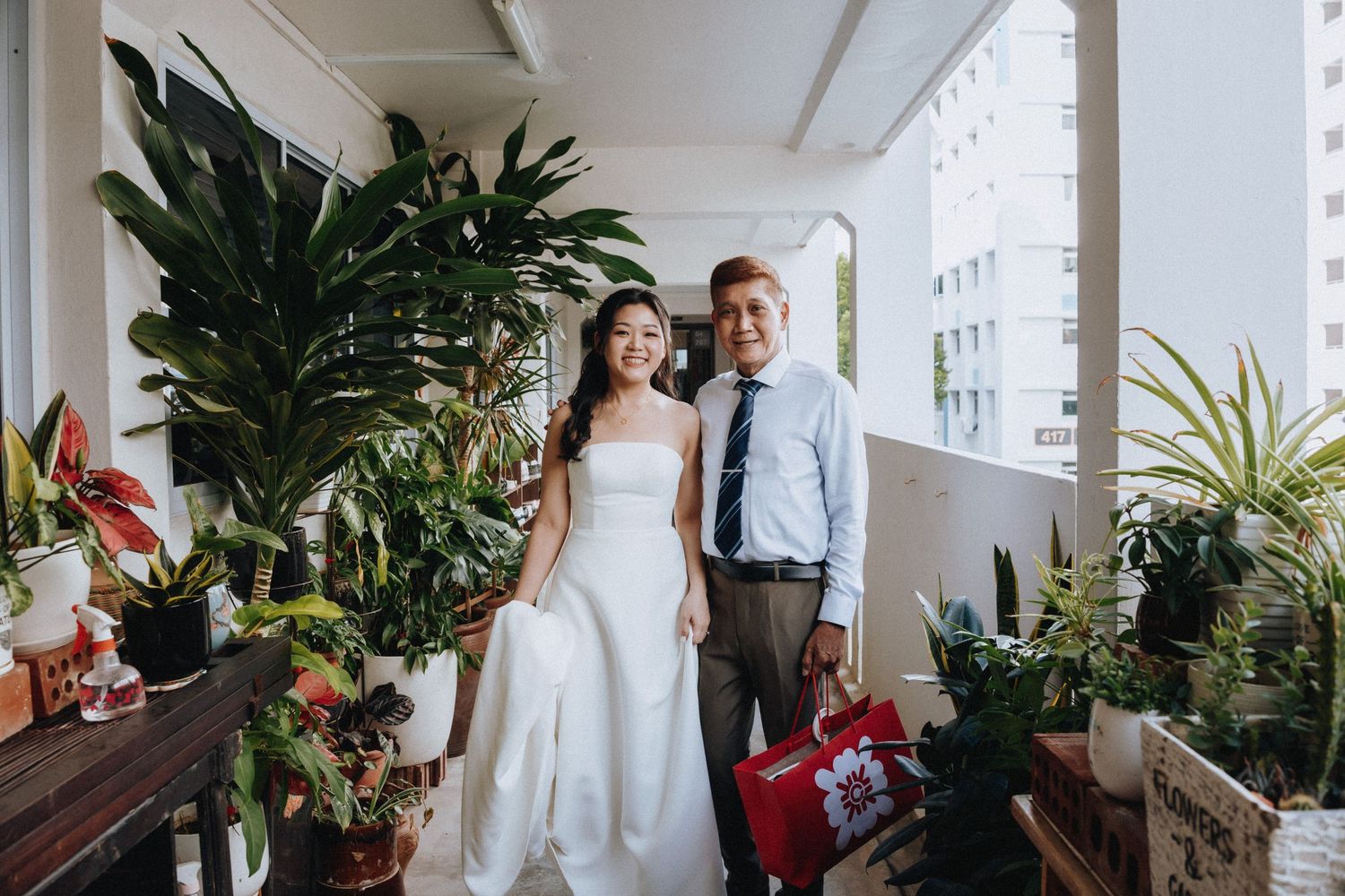 Bride in white gown poses with suited man on plant-filled balcony at wedding celebration.