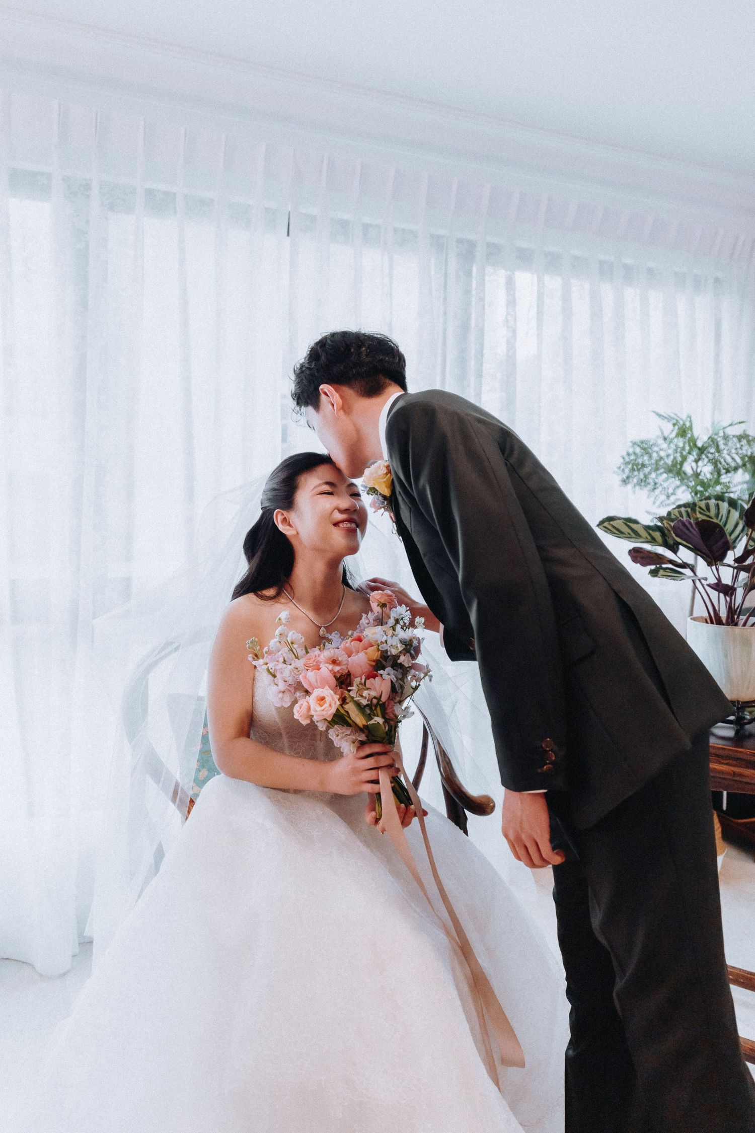 Groom in black suit leaning toward smiling bride in white wedding gown holding colorful bouquet indoors.