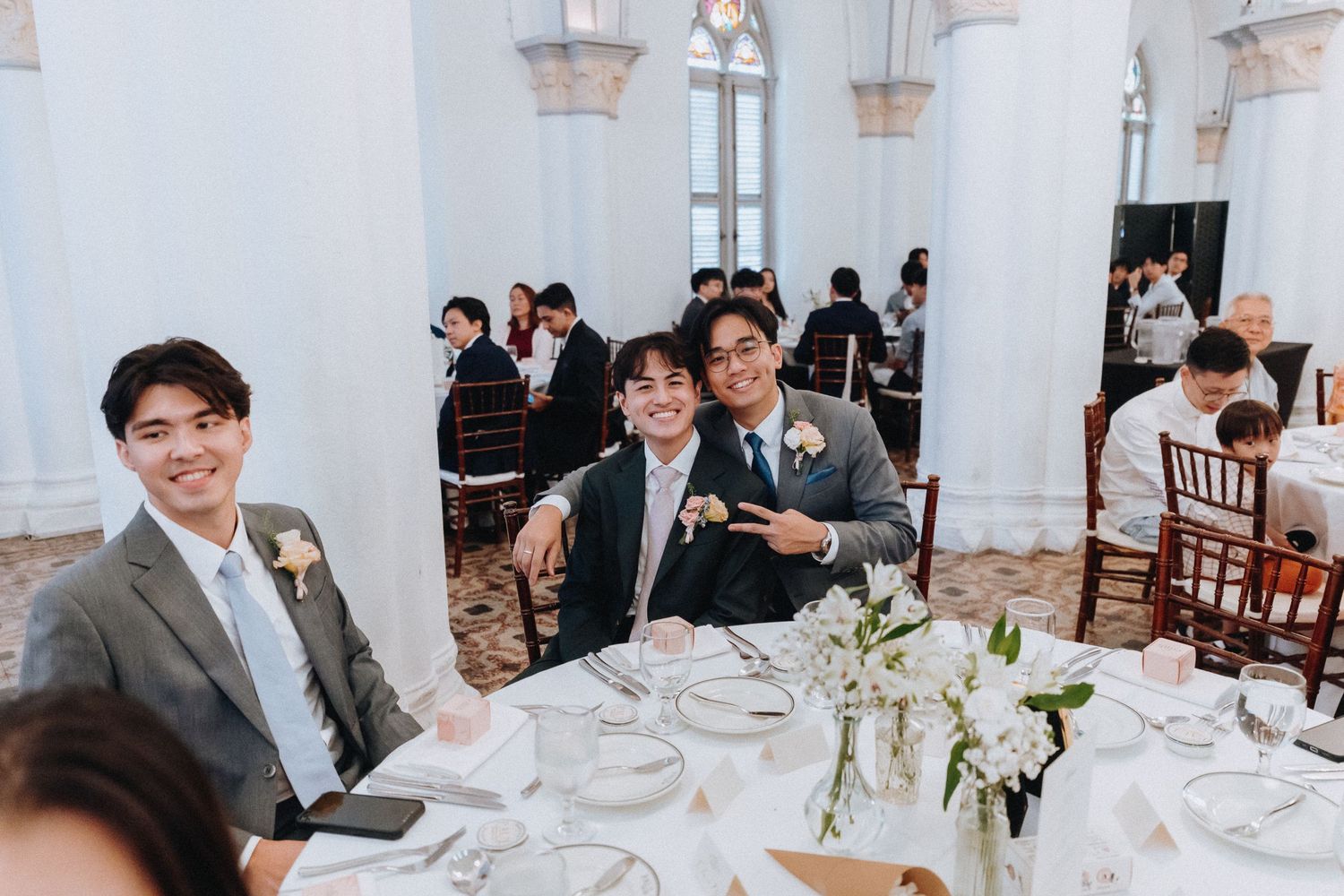 Three young men in suits smile at a wedding reception table in an elegant white ballroom venue.