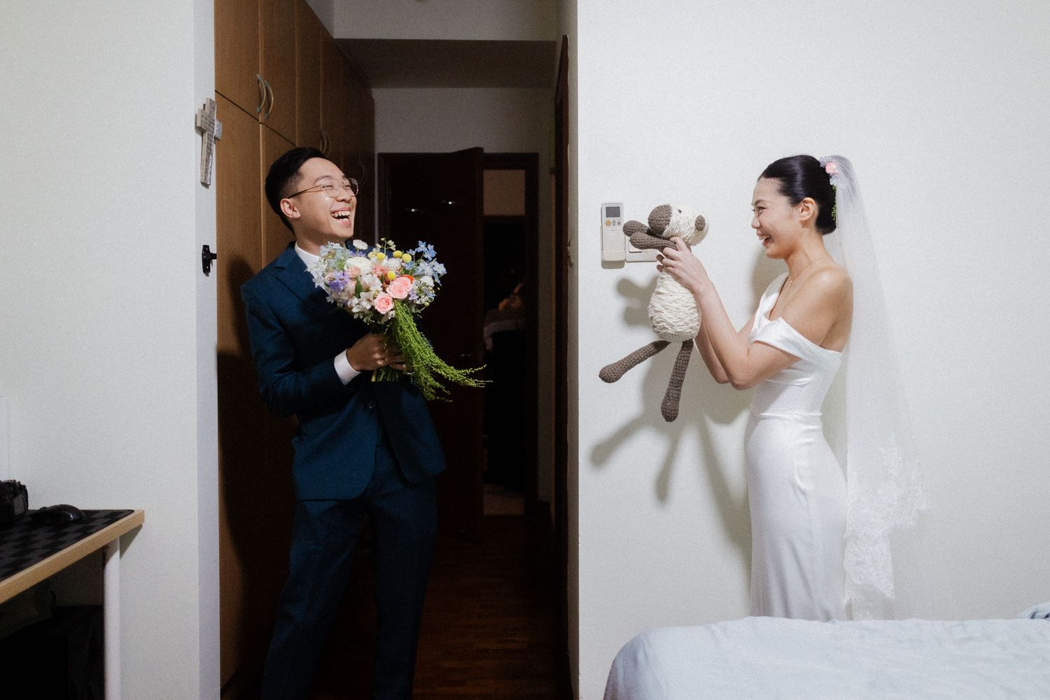 Laughing bride in wedding dress holds stuffed animal toy while groom reacts with joy holding flower bouquet.
