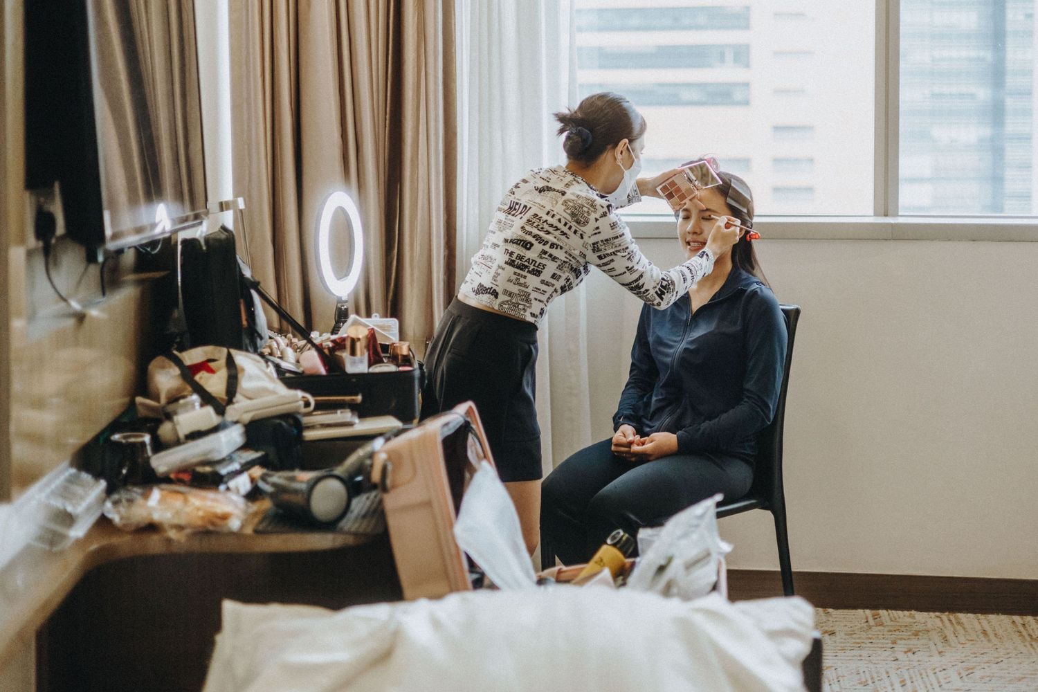 A makeup artist applies makeup to a seated woman in a modern hotel room with a vanity mirror and beauty products nearby.