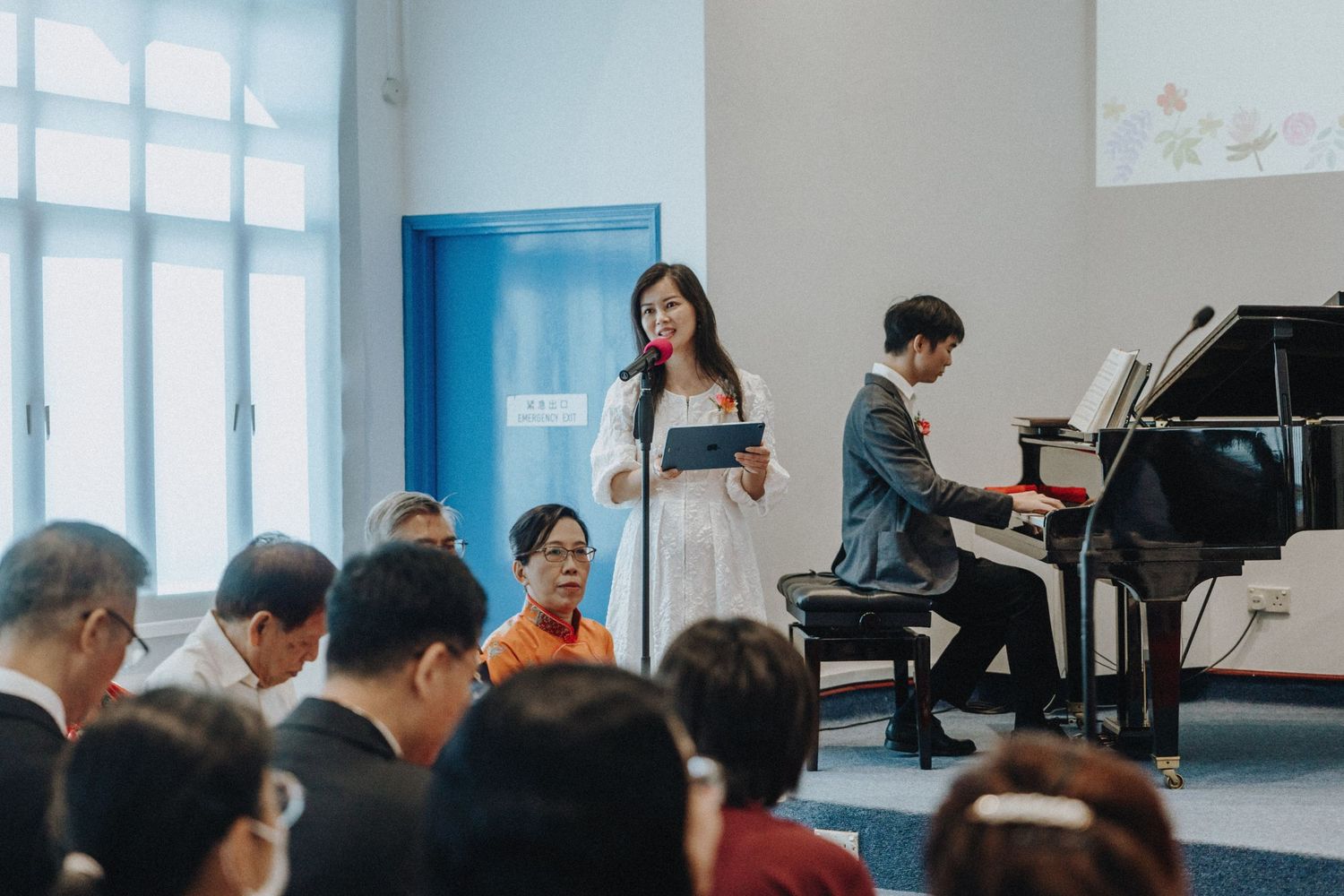A woman in white dress sings at a wedding ceremony while a man plays grand piano for guests.