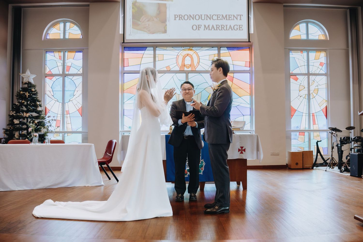A couple exchanges vows during a wedding ceremony with stained glass windows and a Pronouncement of Marriage sign.