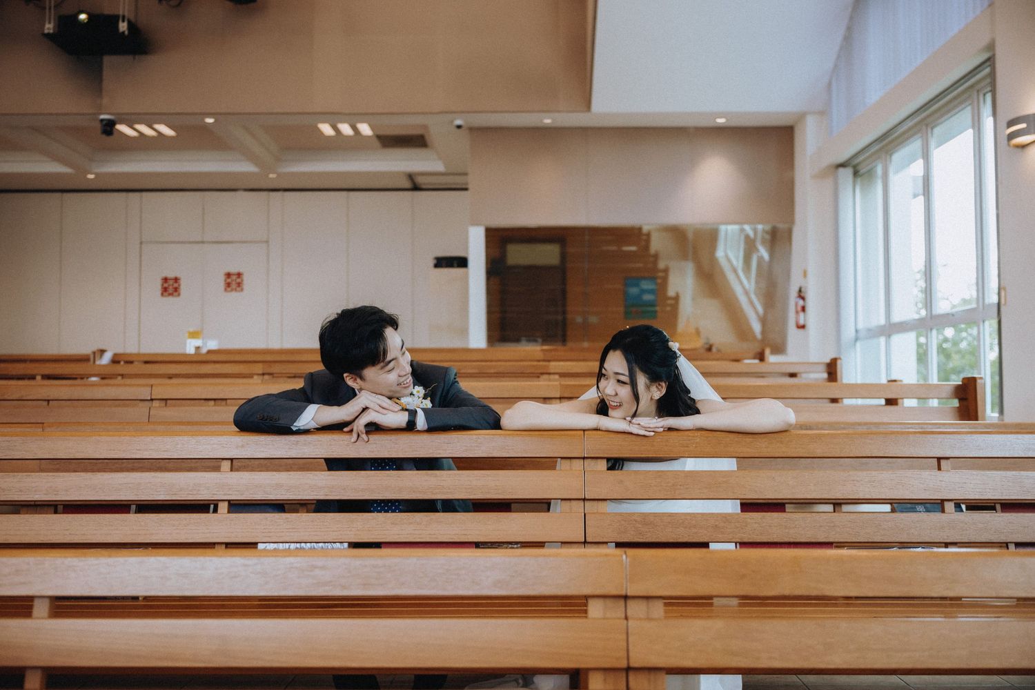 A couple shares a playful moment leaning over wooden church pews during their wedding day.