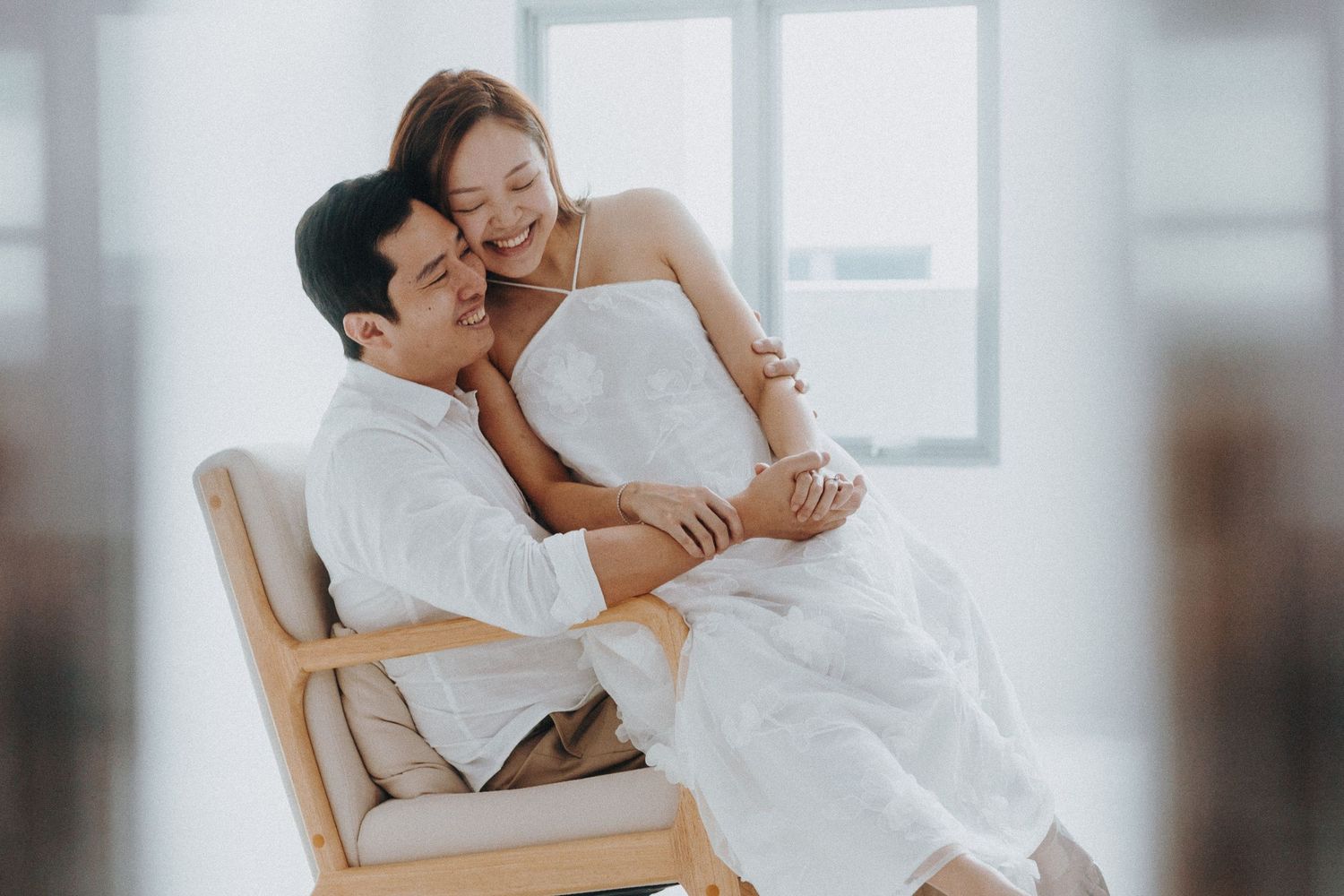 Two people dressed in white sharing a tender moment while sitting in a wooden chair beside a bright window.