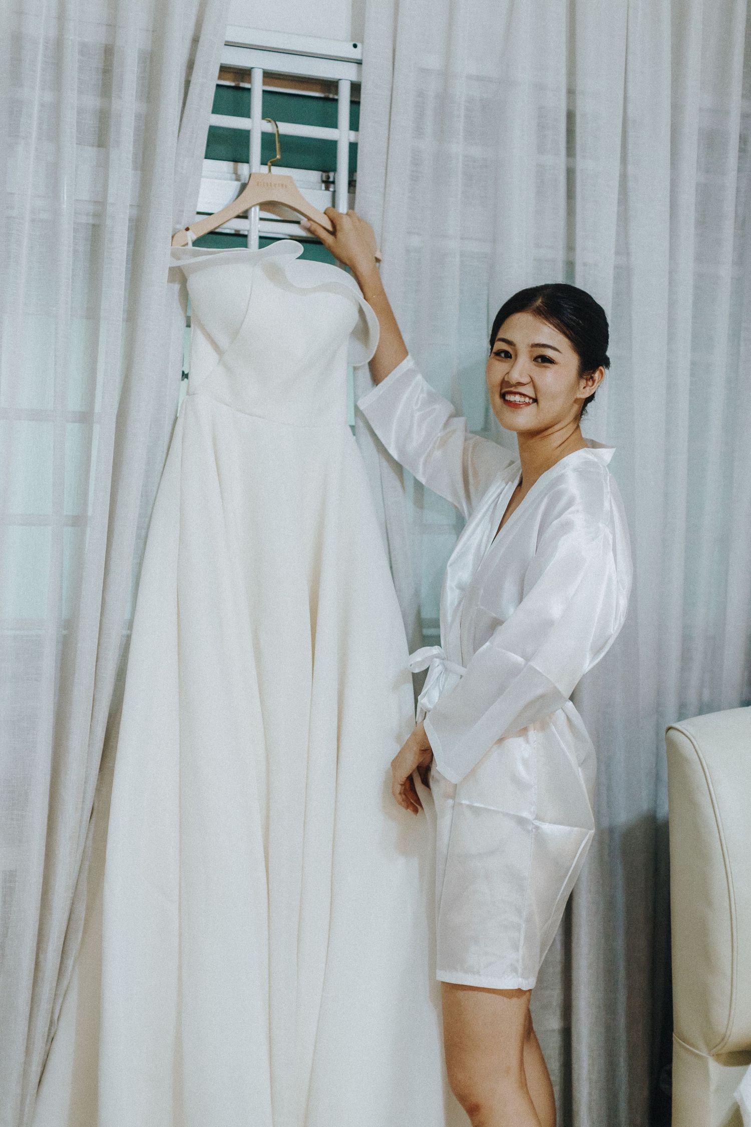 A person in a white silk robe admires a hanging white wedding dress against sheer curtains in a bridal preparation room.
