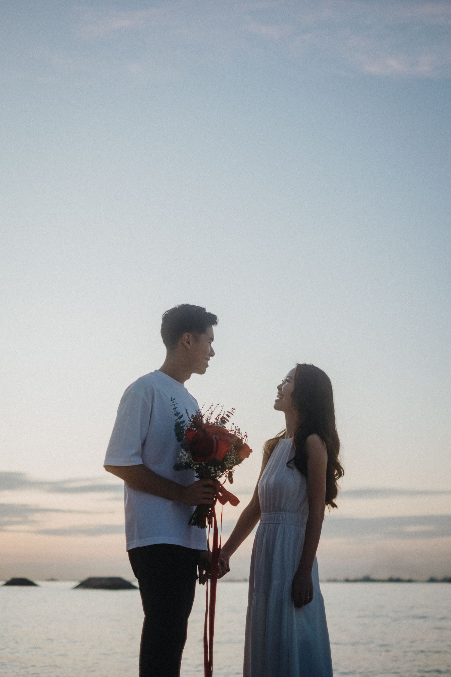 Couple shares a romantic moment by the ocean at sunset with red flowers.