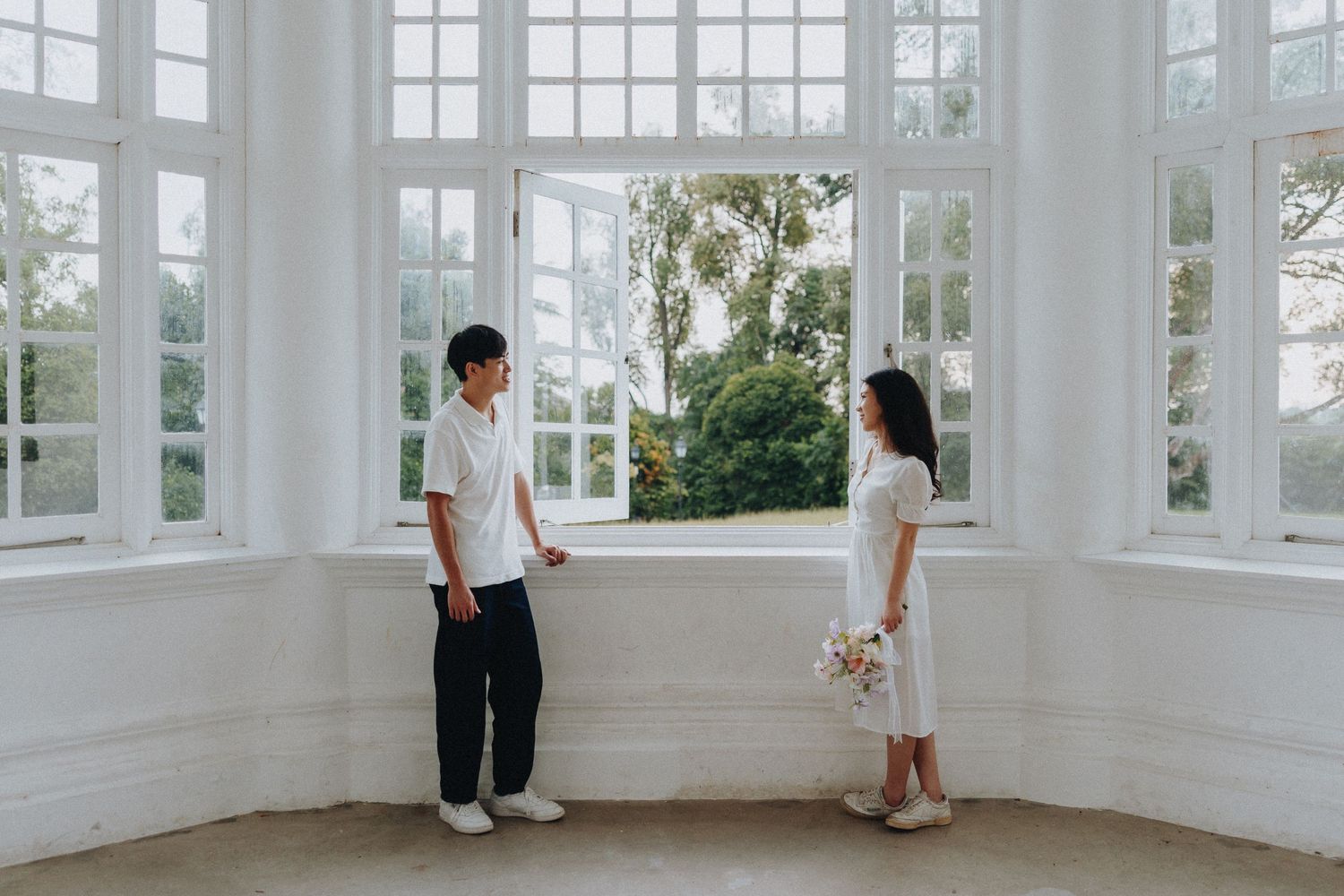 A couple stands in an elegant sunlit white conservatory with tall windows and a garden view.