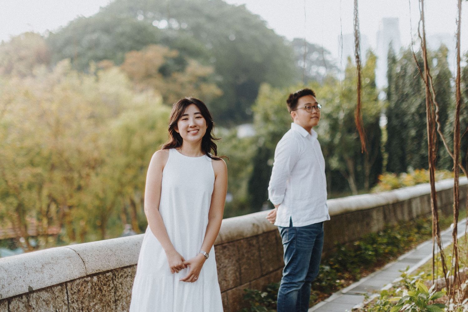 A couple in white wedding attire walks along a scenic mountain path with lush greenery and trees in the distance.