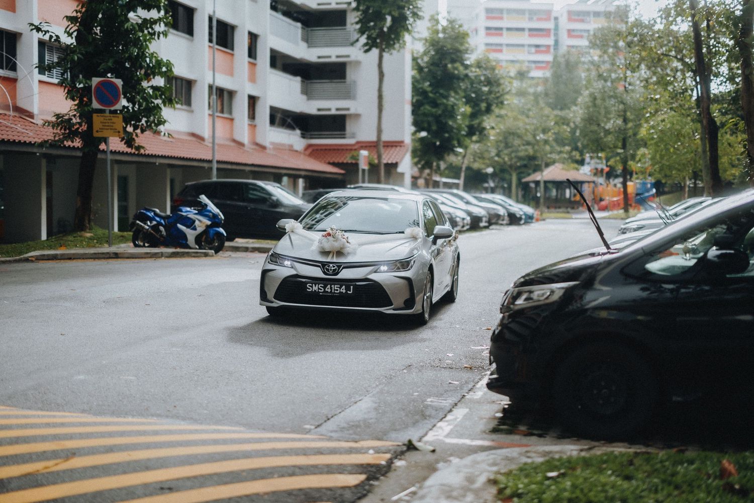 Silver sedan car parked on a wet residential street with apartment buildings in the background.