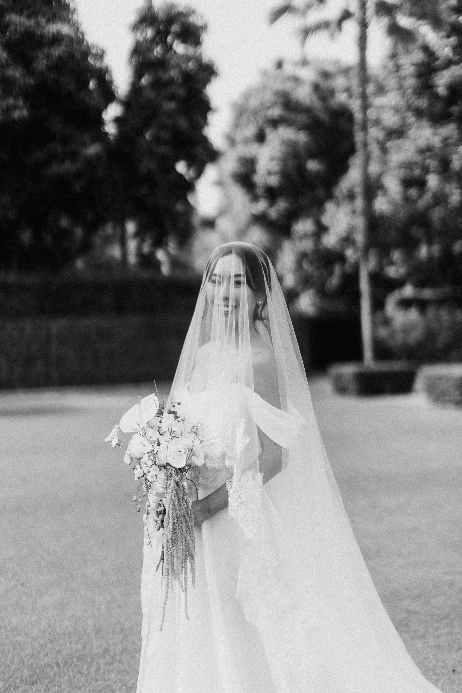 A stunning black and white portrait of a bride in a flowing veil holding an elegant cascading bouquet in a garden setting.