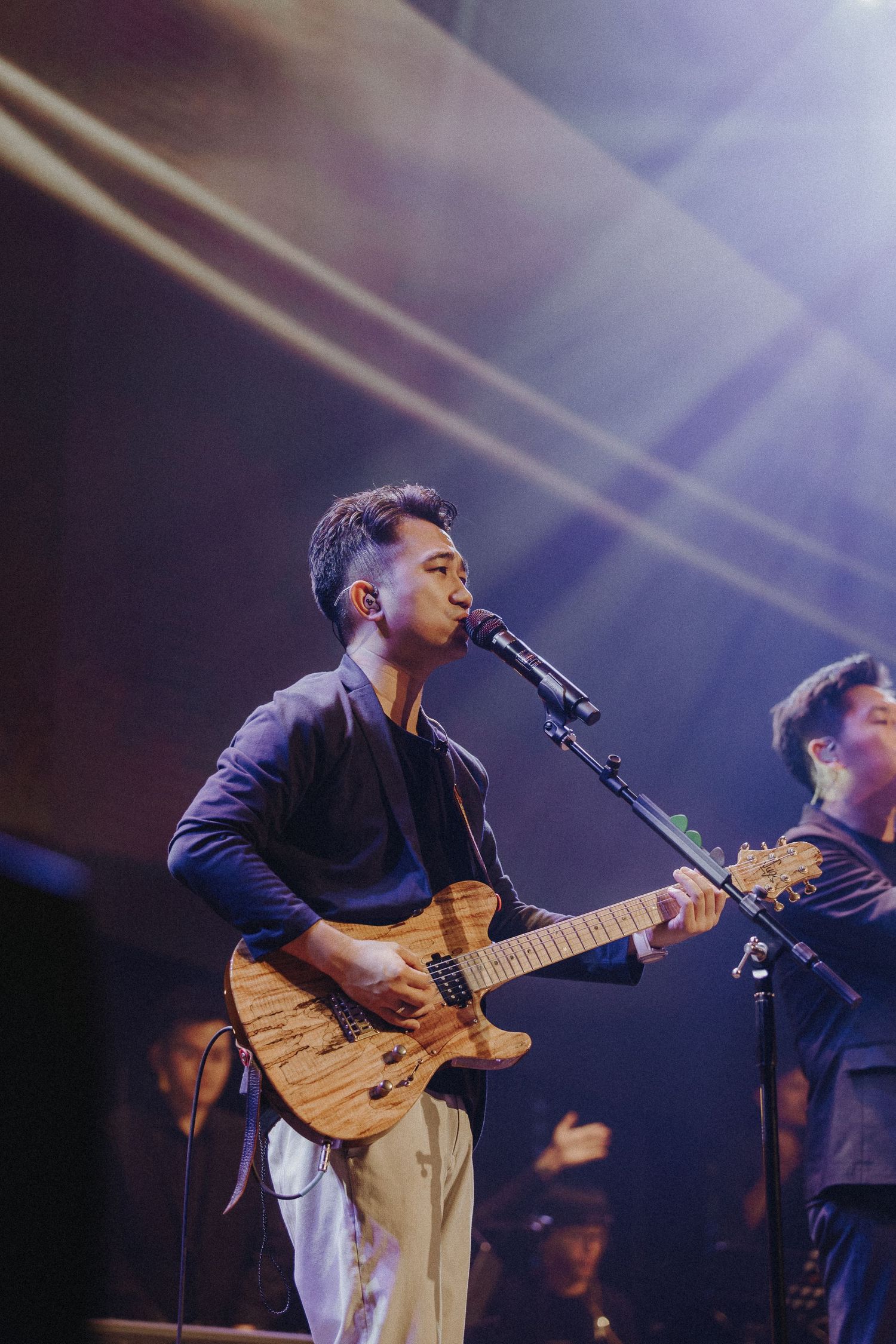 A musician plays an acoustic guitar while singing into a microphone on stage under blue lighting.