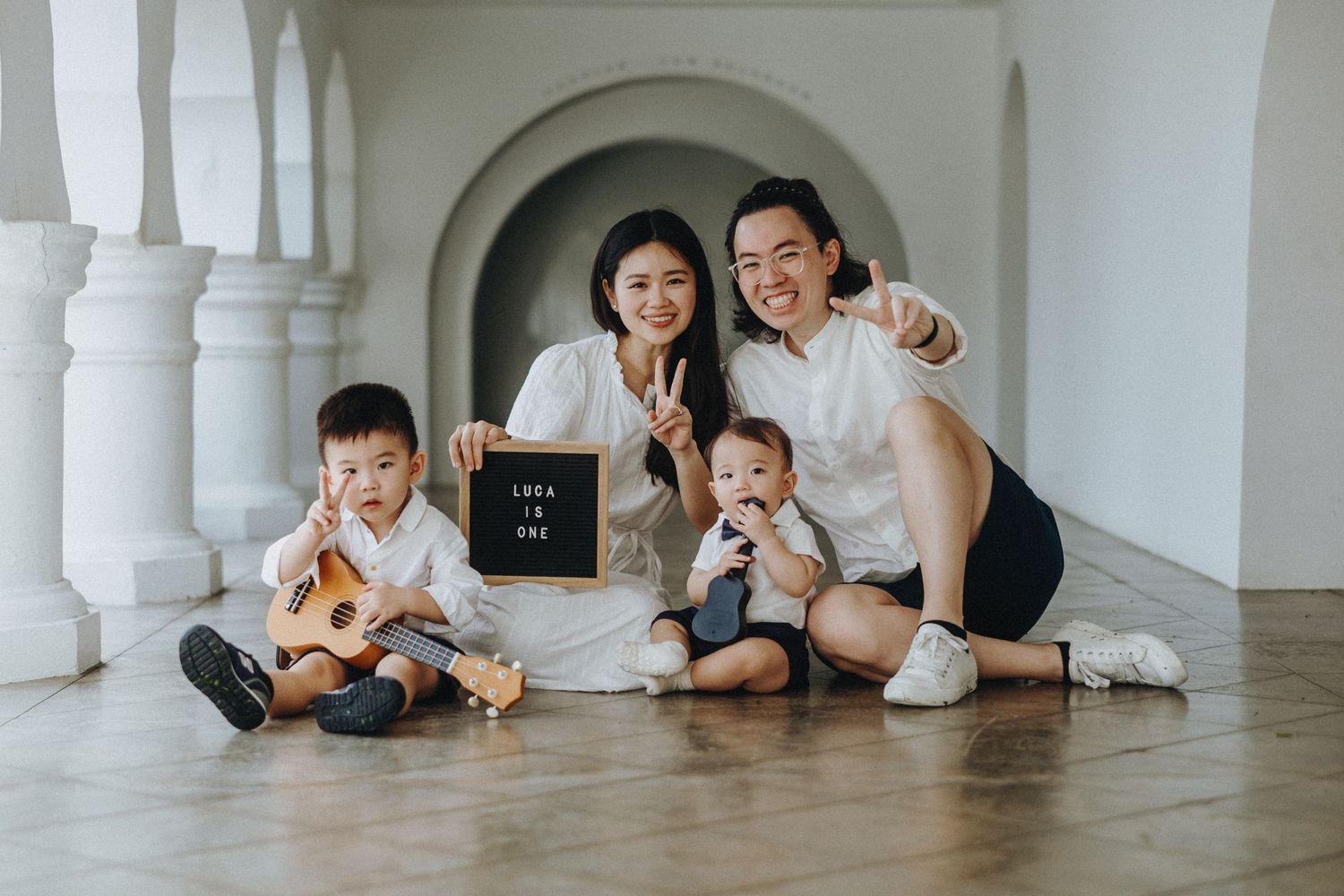 Family dressed in white sitting together on hardwood floor in elegant hallway with white columns holding letter board.