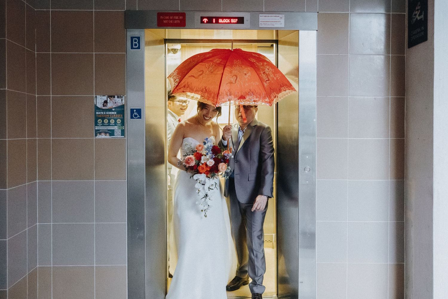 Wedding couple stands in elevator doorway with red umbrella and bouquet.