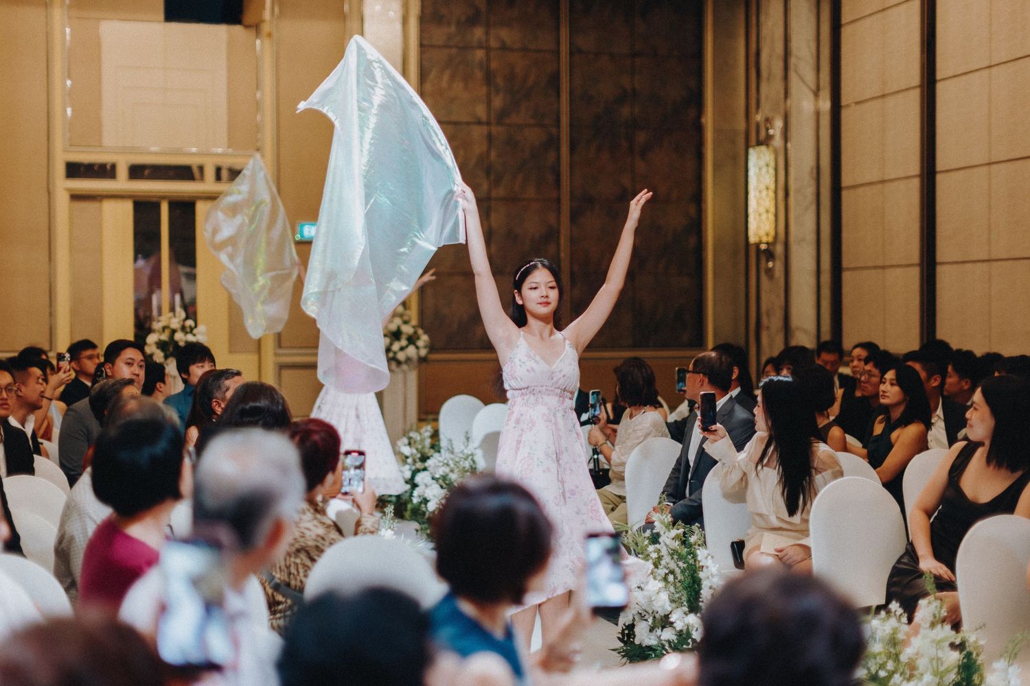 Wedding celebration in a grand ballroom with guests dancing and white floral arrangements.