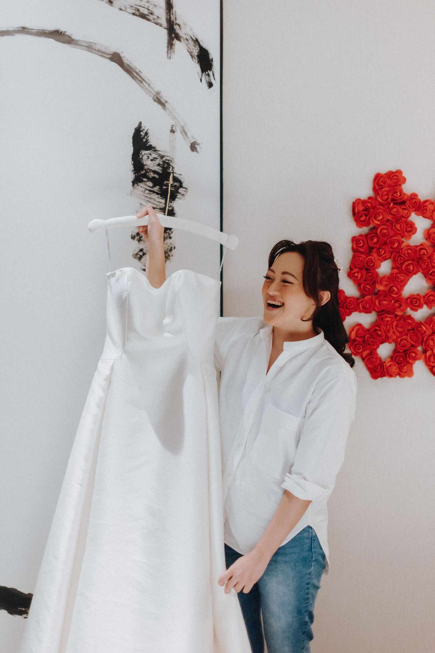 A white wedding dress hangs on display next to decorative red flowers against a light colored wall.