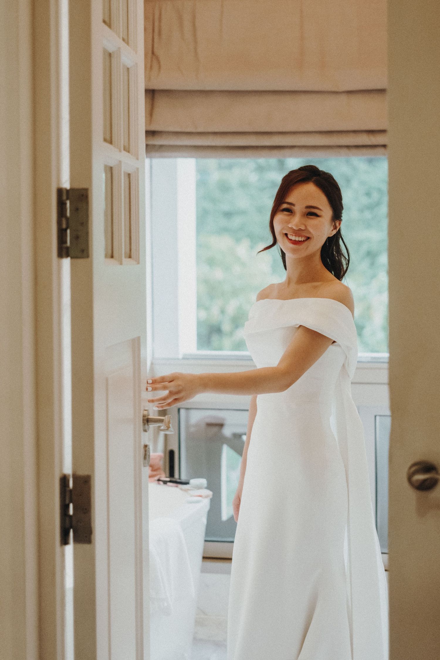 An elegant off-shoulder white wedding dress is shown in a bright interior doorway setting.