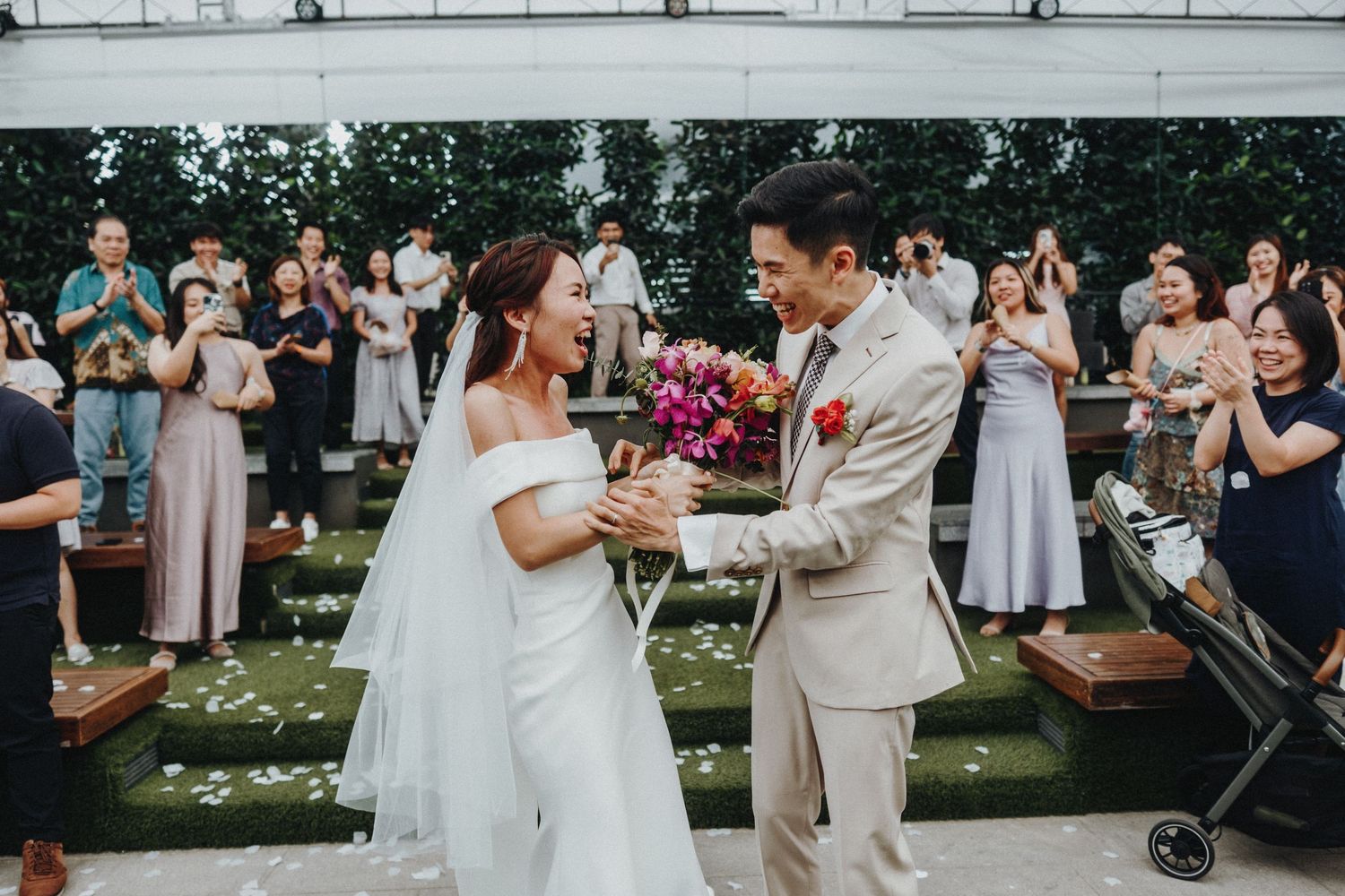 Wedding ceremony outdoors with couple in formal attire exchanging vows on a grass lawn under a floral decorated canopy.