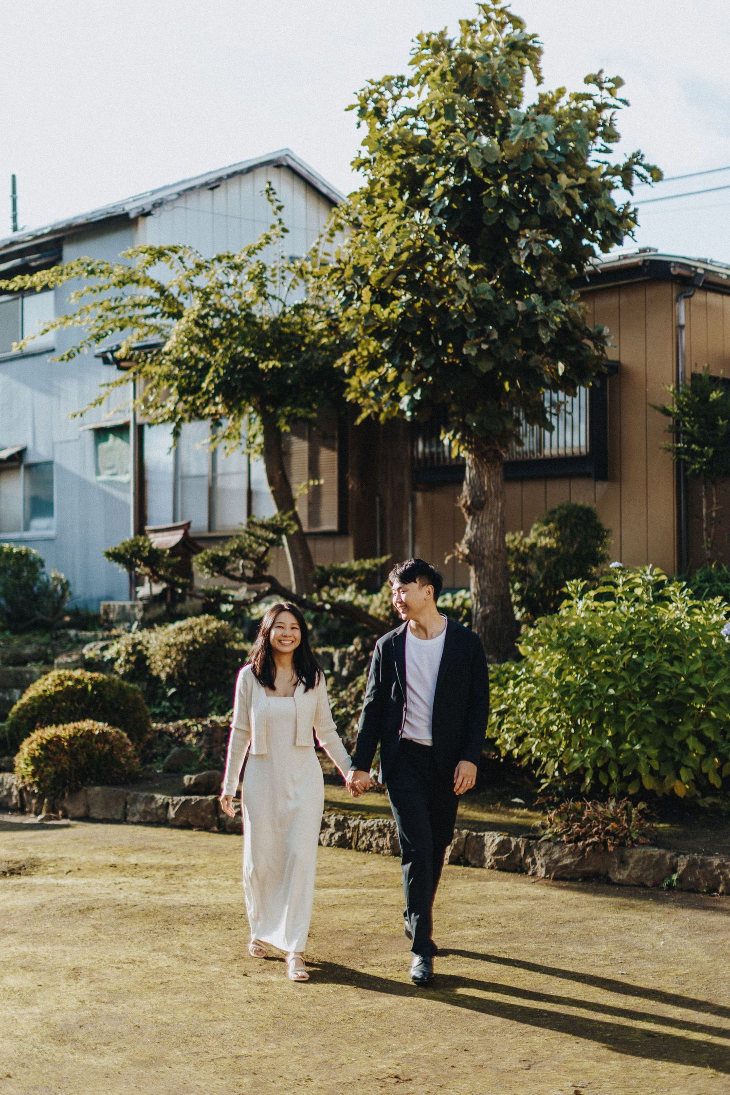 A couple strolling through a sunny garden path with residential buildings visible behind trees.