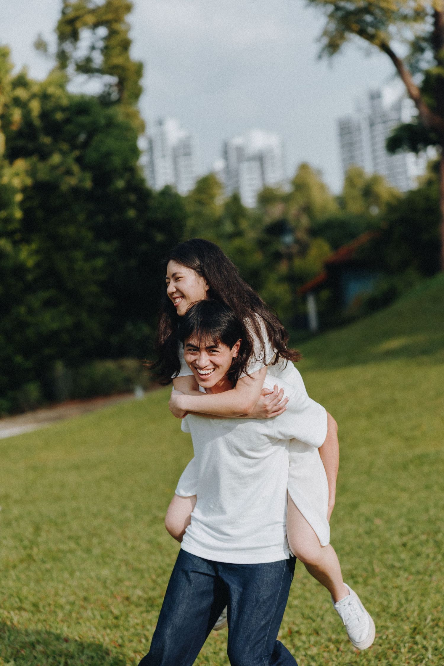 A couple enjoying a playful piggyback ride moment in a park with city buildings visible in the background.