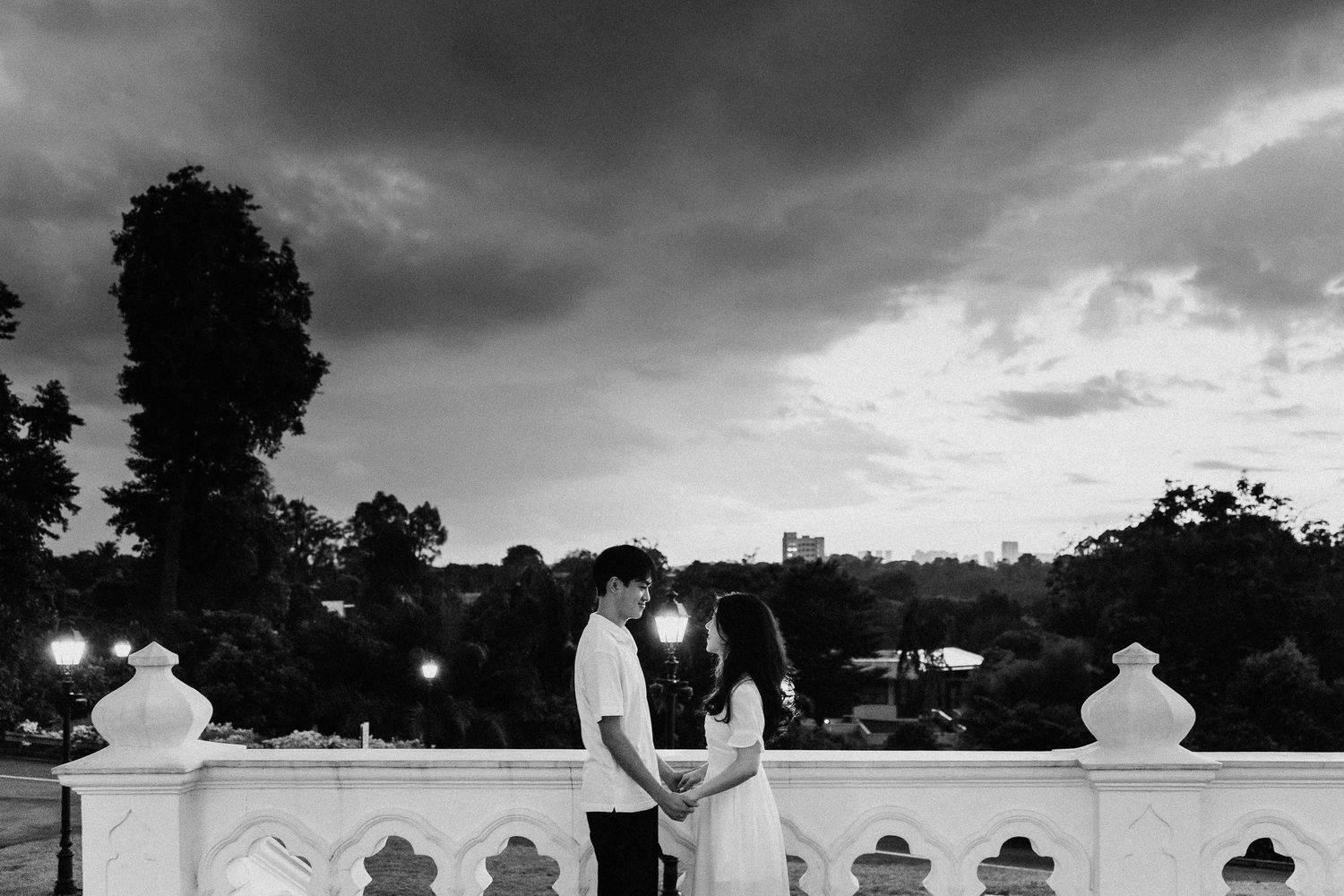 A couple shares an intimate moment on a white stone balcony under dramatic storm clouds in black and white.