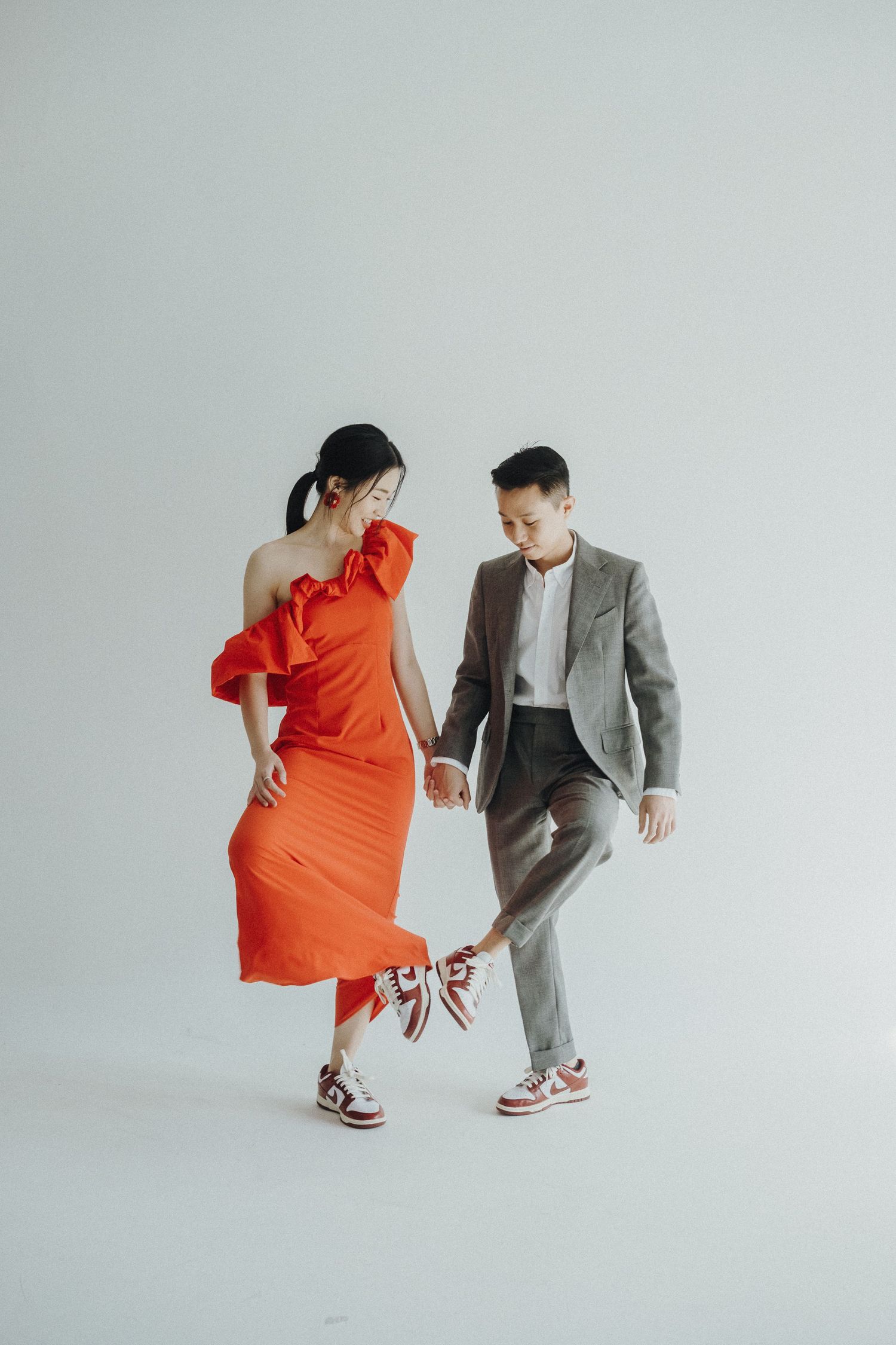Stylish couple in orange dress and gray suit dancing and posing against white background.