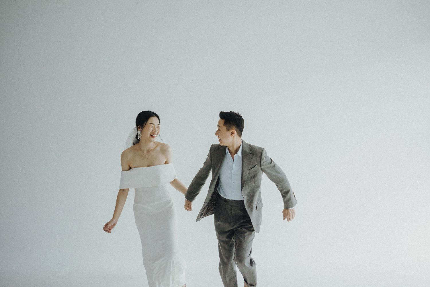 Bride and groom playfully hold hands while dancing joyfully across a white studio backdrop at their wedding photoshoot.