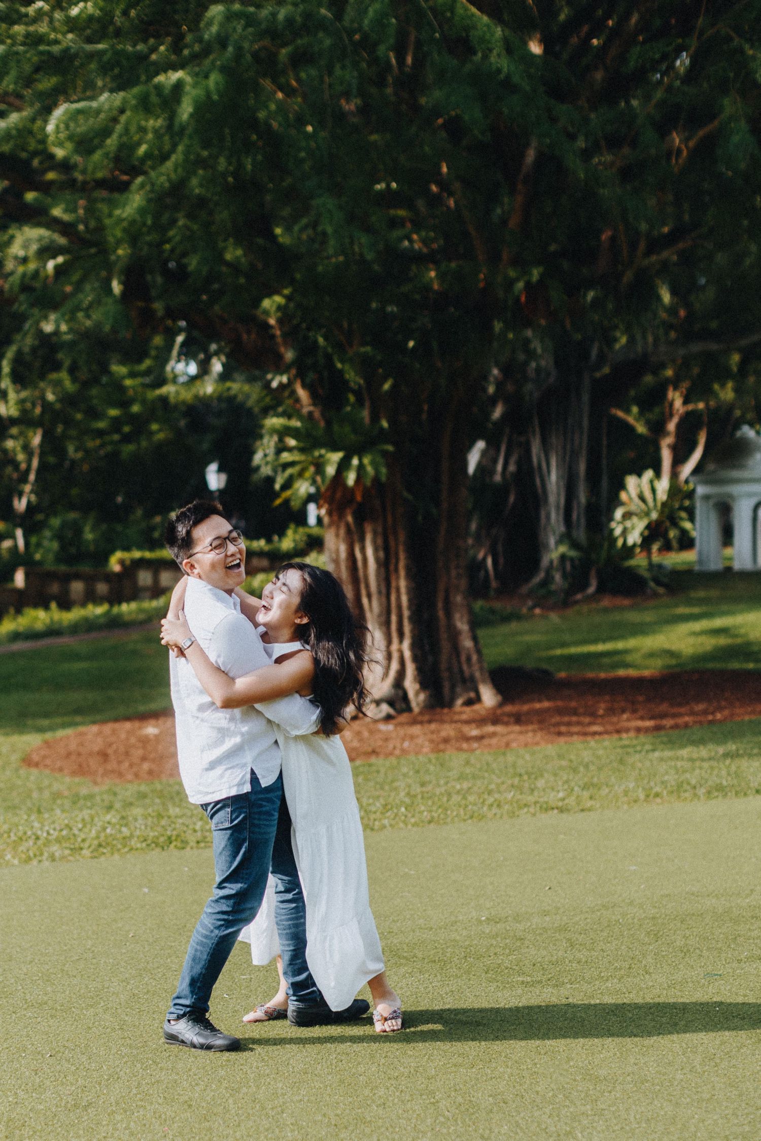 A romantic couple embraces while sharing a laughing moment on a sunlit lawn with large trees in the background.