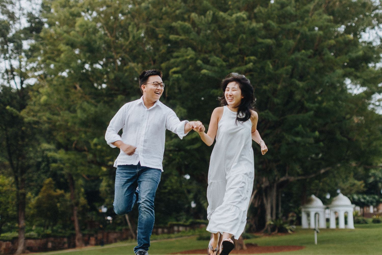 Joyful couple running playfully together on green lawn with trees and white house in background.